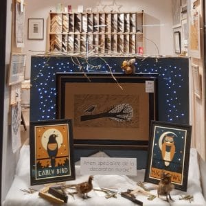 Vitrine de Noël d'Artem avec quelques affiches seules présentées sur des cintres pantalon et autres affiches encadrées, sur les affiches des oiseaux, des chouettes, des animaux pour décorer : un écureuil, un faisan, une chouette, quelques échantillons de baguettes et de passe-partout. En arrière plan le présentoir de baguettes en bois et en aluminium.