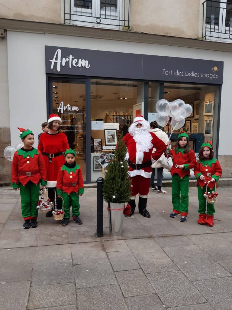 Mère Noël, Père Noël et leurs lutins sont devant la boutique Artem au 16 rue Franklin à Nantes. Ils offrent des ballons, des bonbons et des chocolats pour fêter Noël. Il y a un sapin de Noël dans un pot métallique avec un nœud rouge.